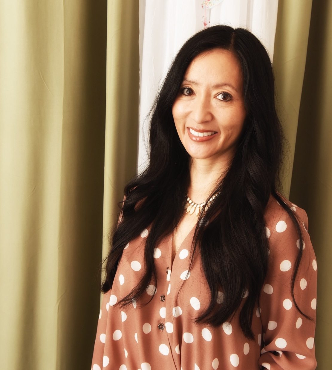 Lorin's profile picture in pink and white polka dot shirt standing next curtains in office
